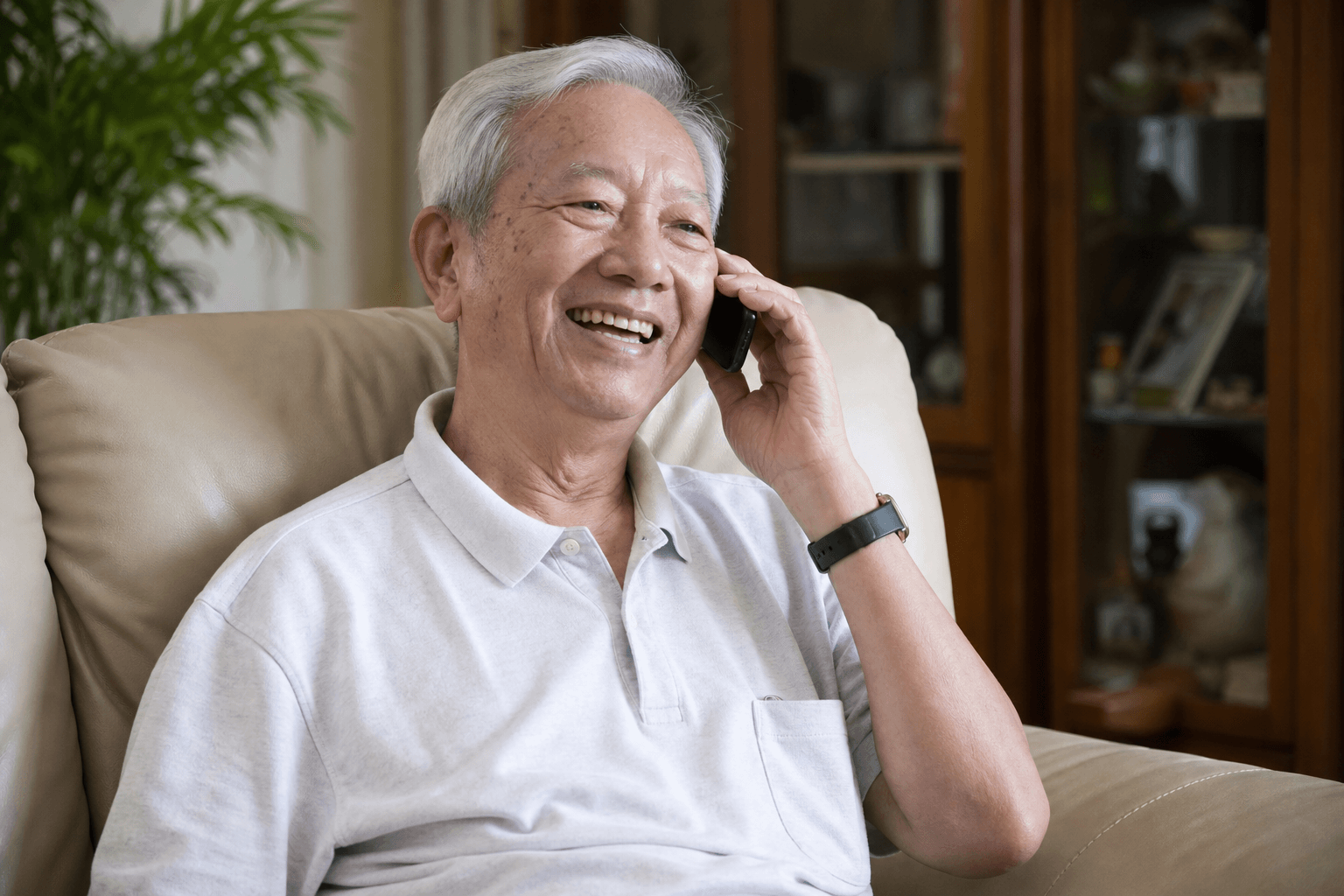 Elderly Singaporean man smiling while talking on a smartphone in a bright living room